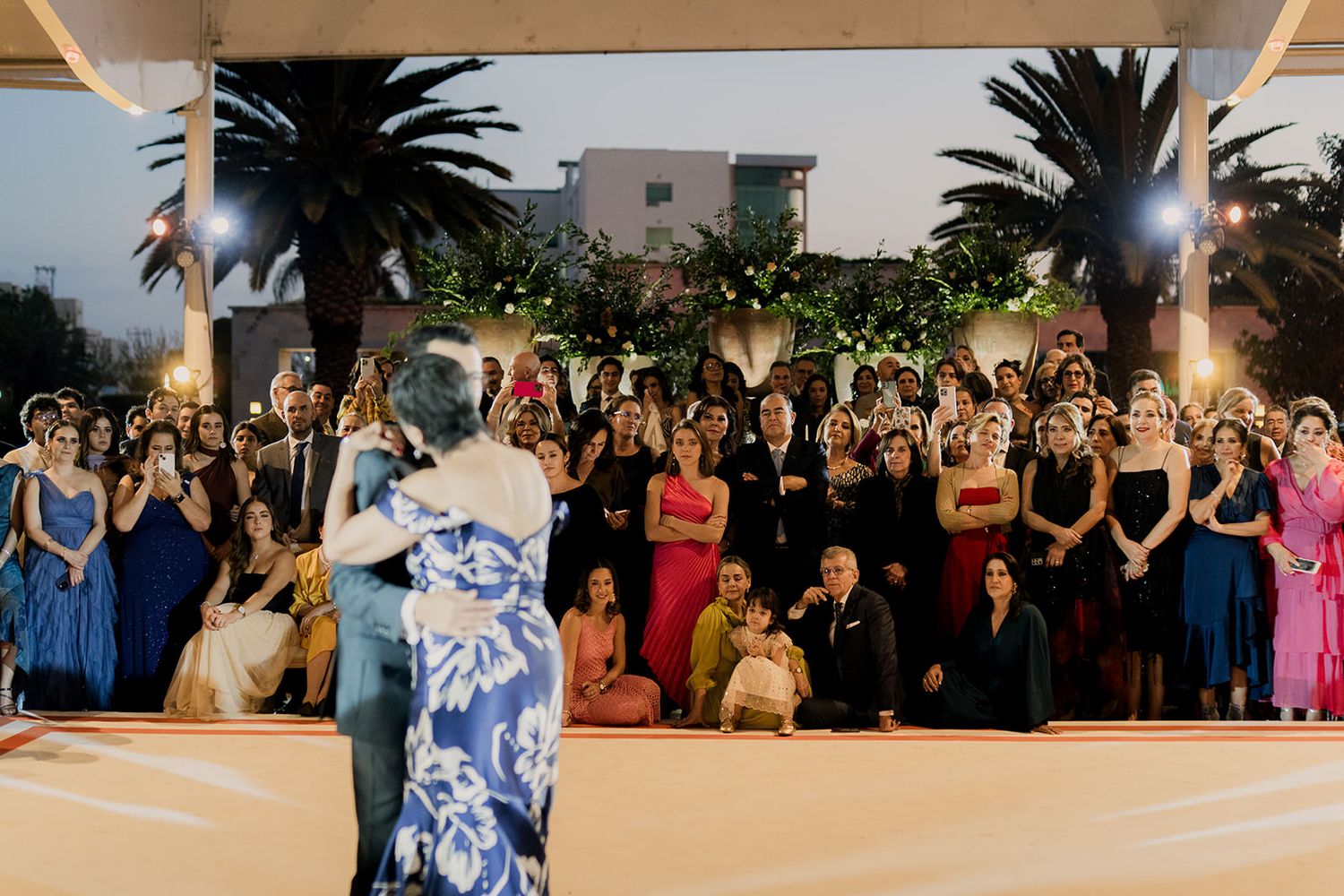 Couple shares first dance at outdoor wedding reception as guests watch under palm trees and warm evening lights.