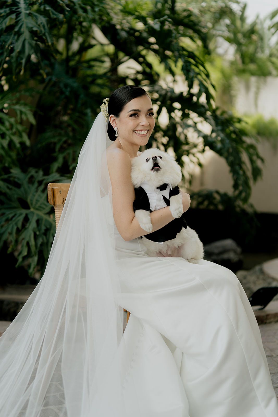 Smiling bride in white gown and veil holding a small white dog dressed in a tuxedo at an outdoor wedding.