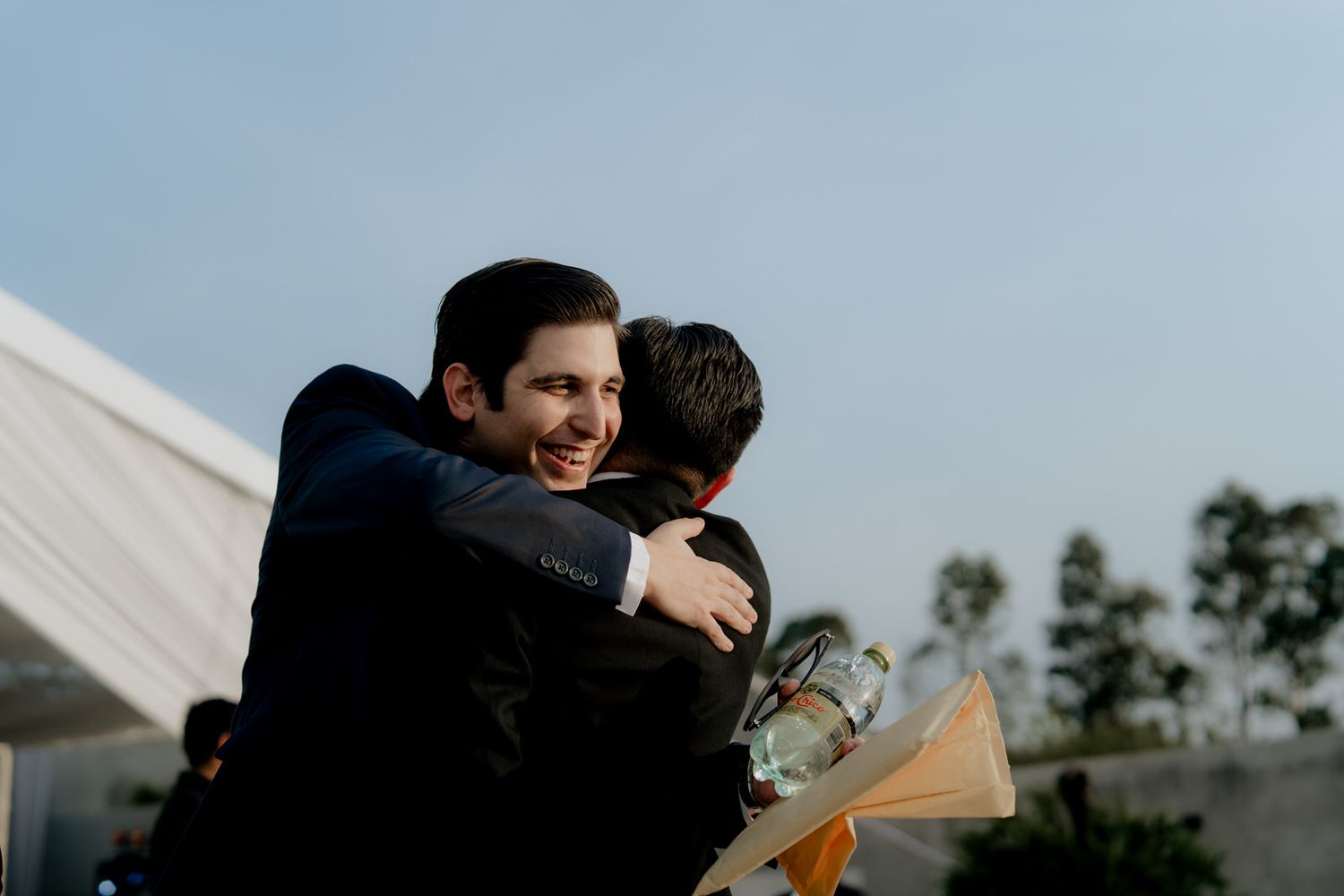 Two men sharing a warm hug at an outdoor wedding celebration under a clear blue sky.