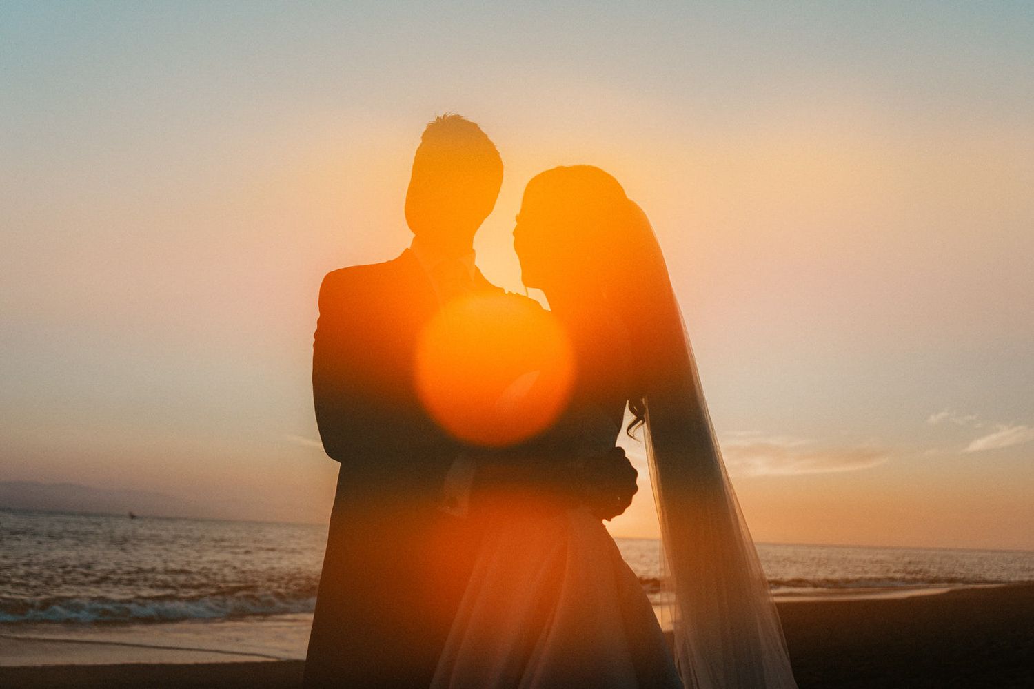 Silhouettes embrace against a dramatic orange sunset sky over the ocean.