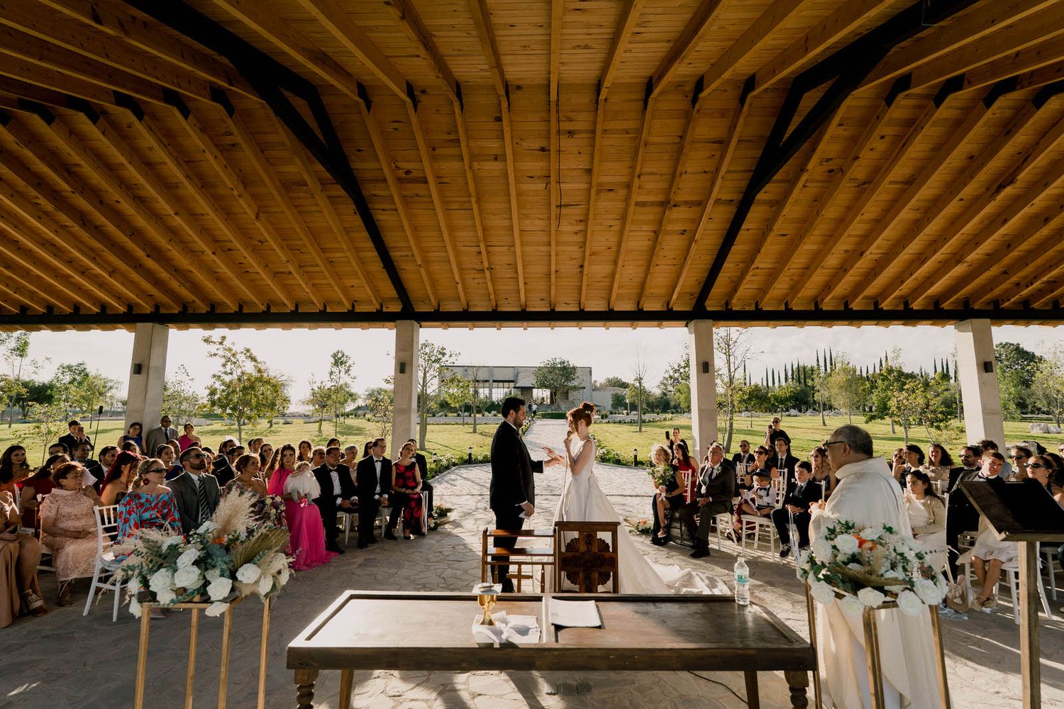 Wide angle view of wedding ceremony under wooden beamed pavilion roof.