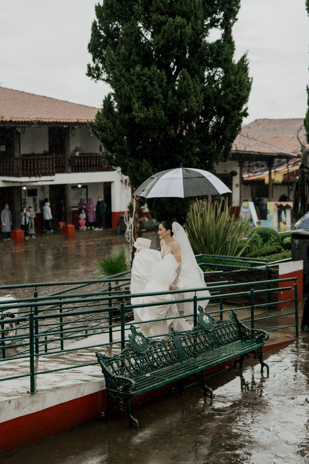 Wedding scene on a rainy day with umbrellas and colonial buildings in background.