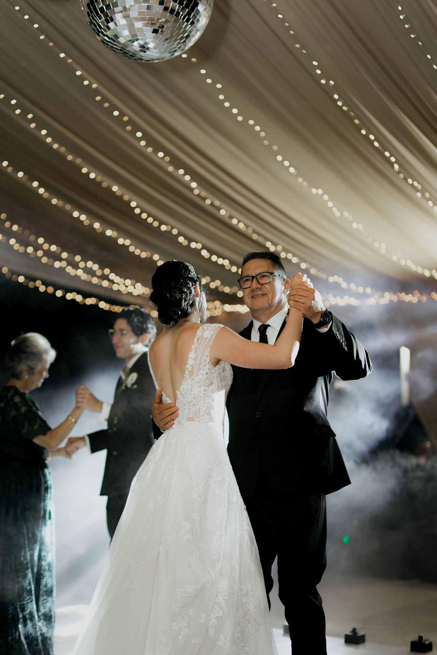 Couples slow dancing beneath elegant ceiling draping and string lights at a wedding celebration.
