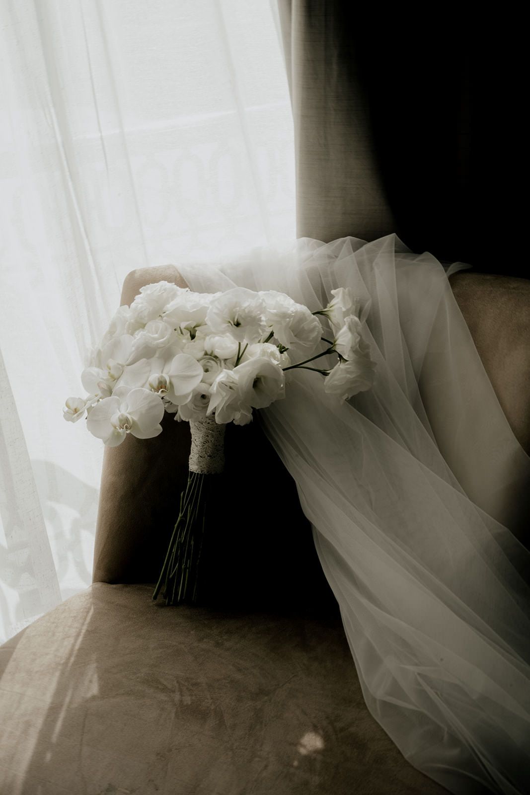 Elegant white rose bridal bouquet resting on a velvet chair with a flowing veil beside sheer curtains.