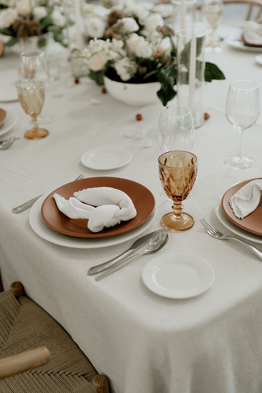 Elegant wedding table setting with terracotta chargers, amber goblets, white napkins, and floral centerpiece.