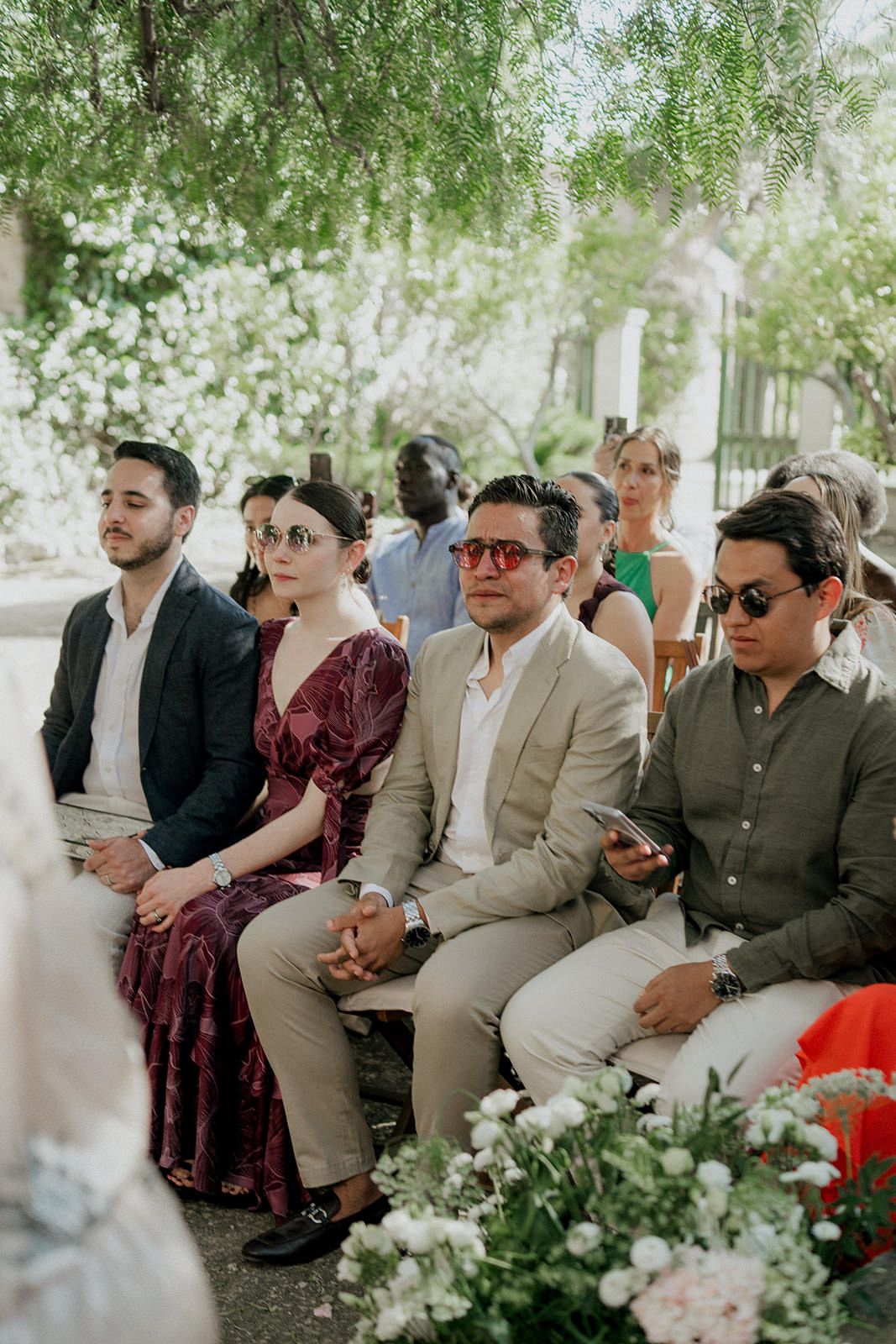 Wedding guests seated outdoors under trees watching ceremony with floral decorations in foreground.
