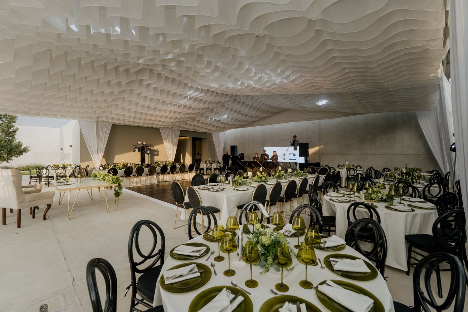 Elegant banquet hall with sculptural white ceiling, round tables with green floral centerpieces, and black chairs for a formal event.