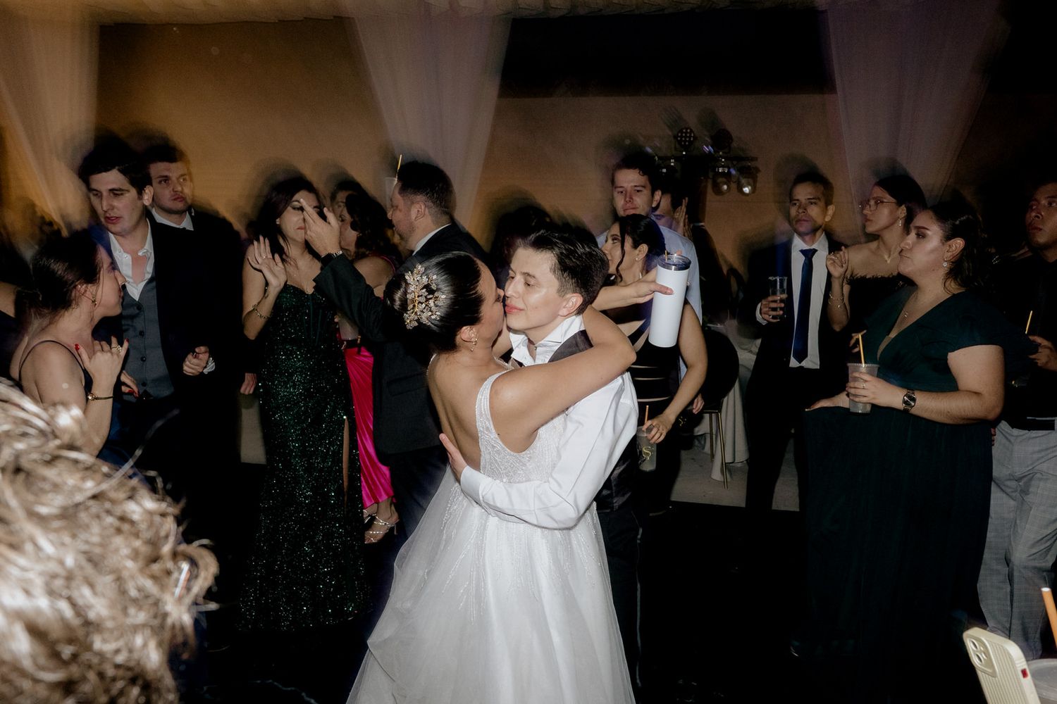 Bride and groom share a first dance surrounded by elegantly dressed wedding guests at a lively indoor reception.