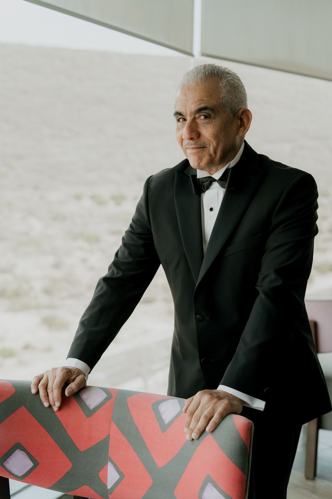 Elegant older man in black tuxedo with bow tie leaning on colorful chair.