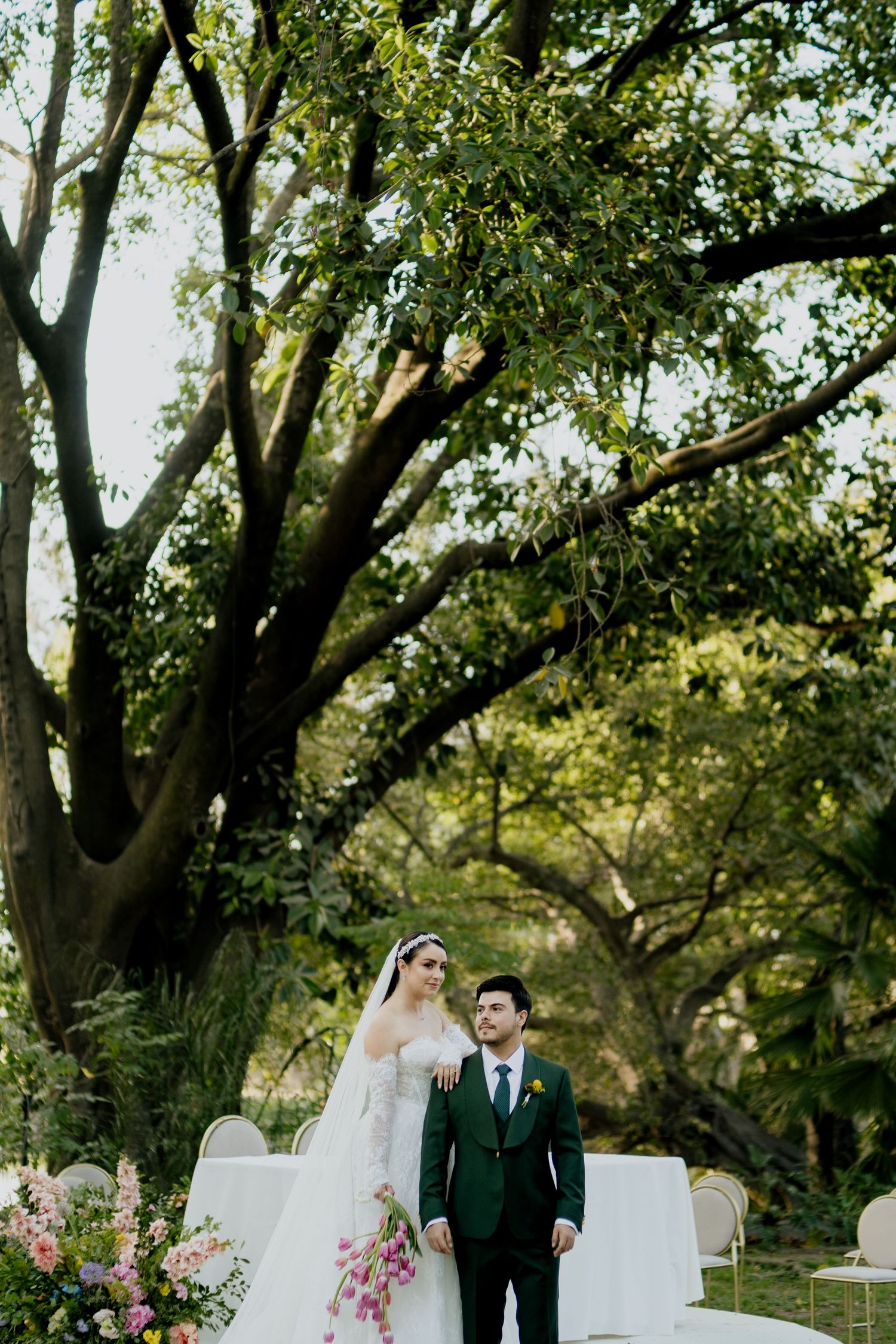 Bride and groom stand together outdoors under a large tree during an elegant garden wedding ceremony with floral arrangements.