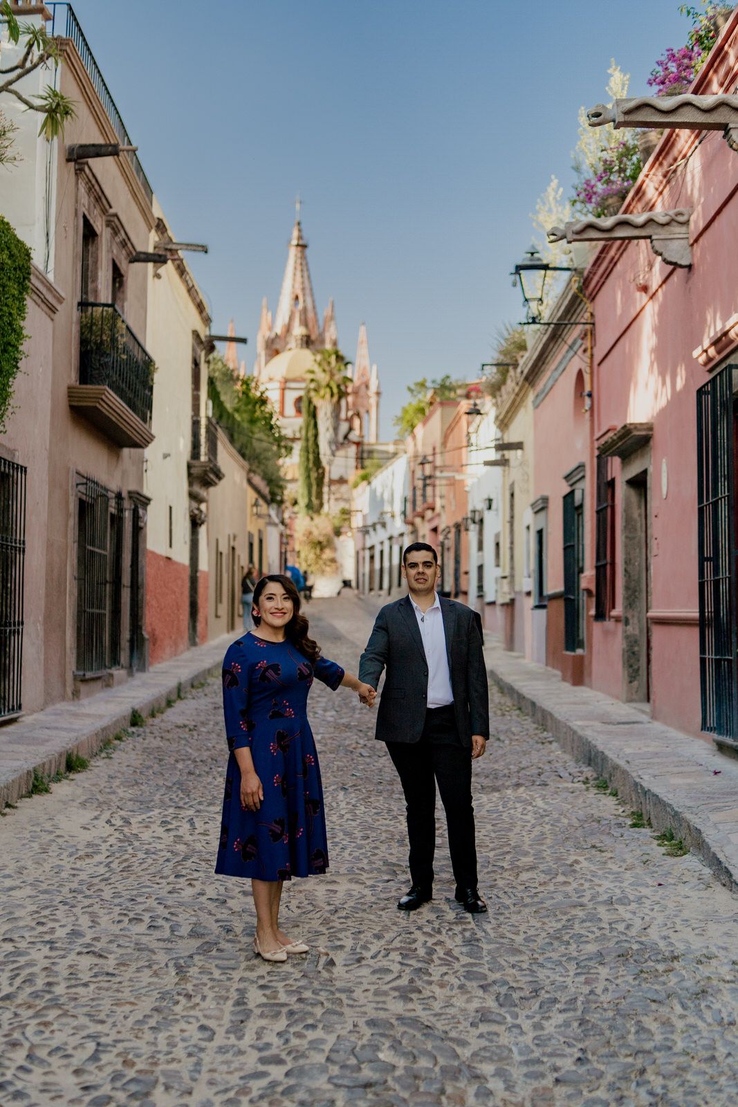 Couple poses together on a charming cobblestone street lined with pink and white colonial buildings.