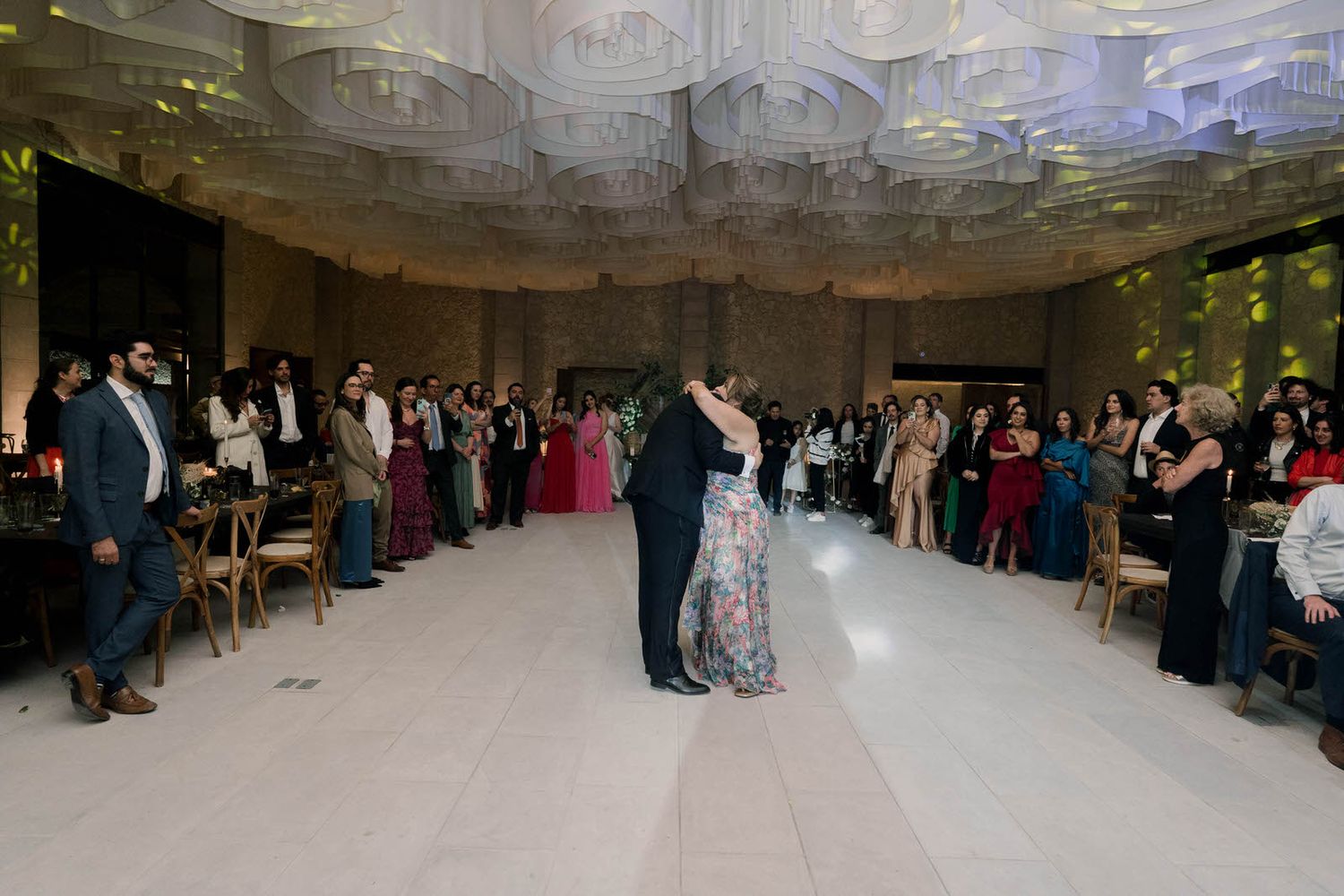 Wedding guests gather on a dance floor in an elegant indoor venue with decorative ceiling details.