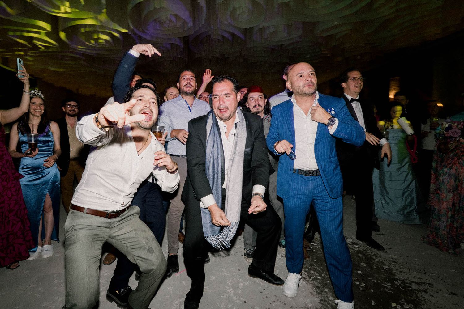 Enthusiastic wedding guests dance and celebrate on a dimly lit dance floor.