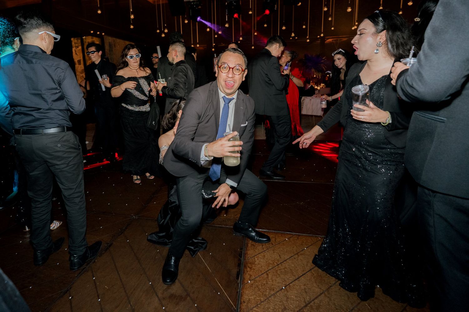Partygoers dancing and celebrating on a dark dance floor with ambient red lighting.