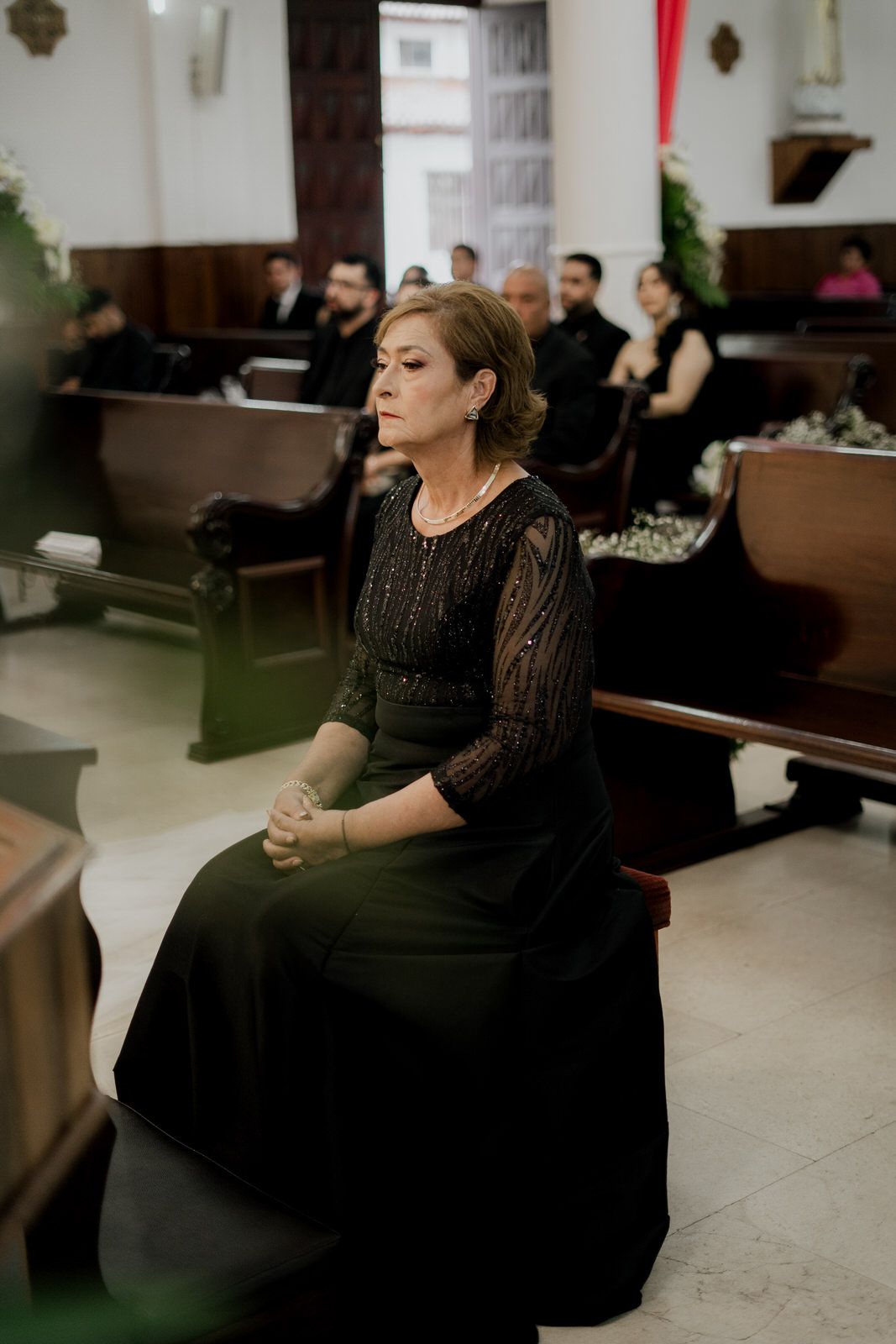 A guest in an elegant black dress sits thoughtfully in a church pew during a ceremony.