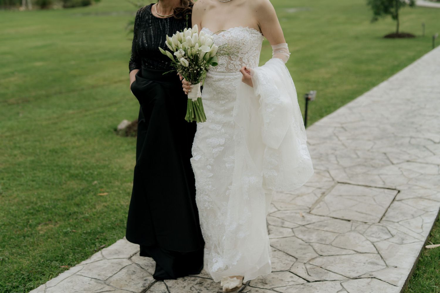 Newlyweds stroll along rustic stone pathway with lush green grass in background.