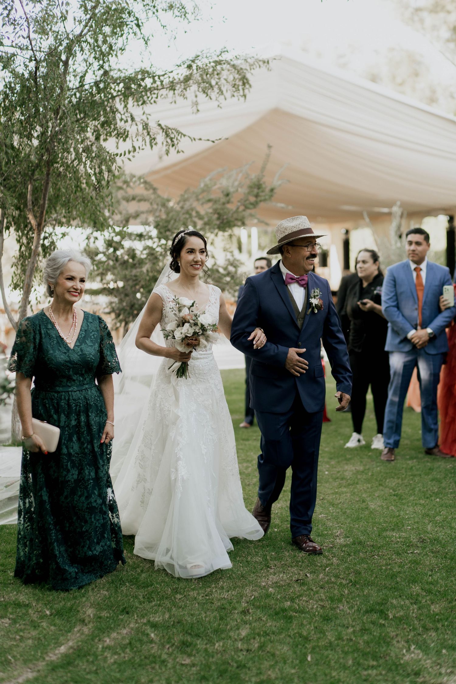 Wedding party walks through garden setting under elegant white tent.