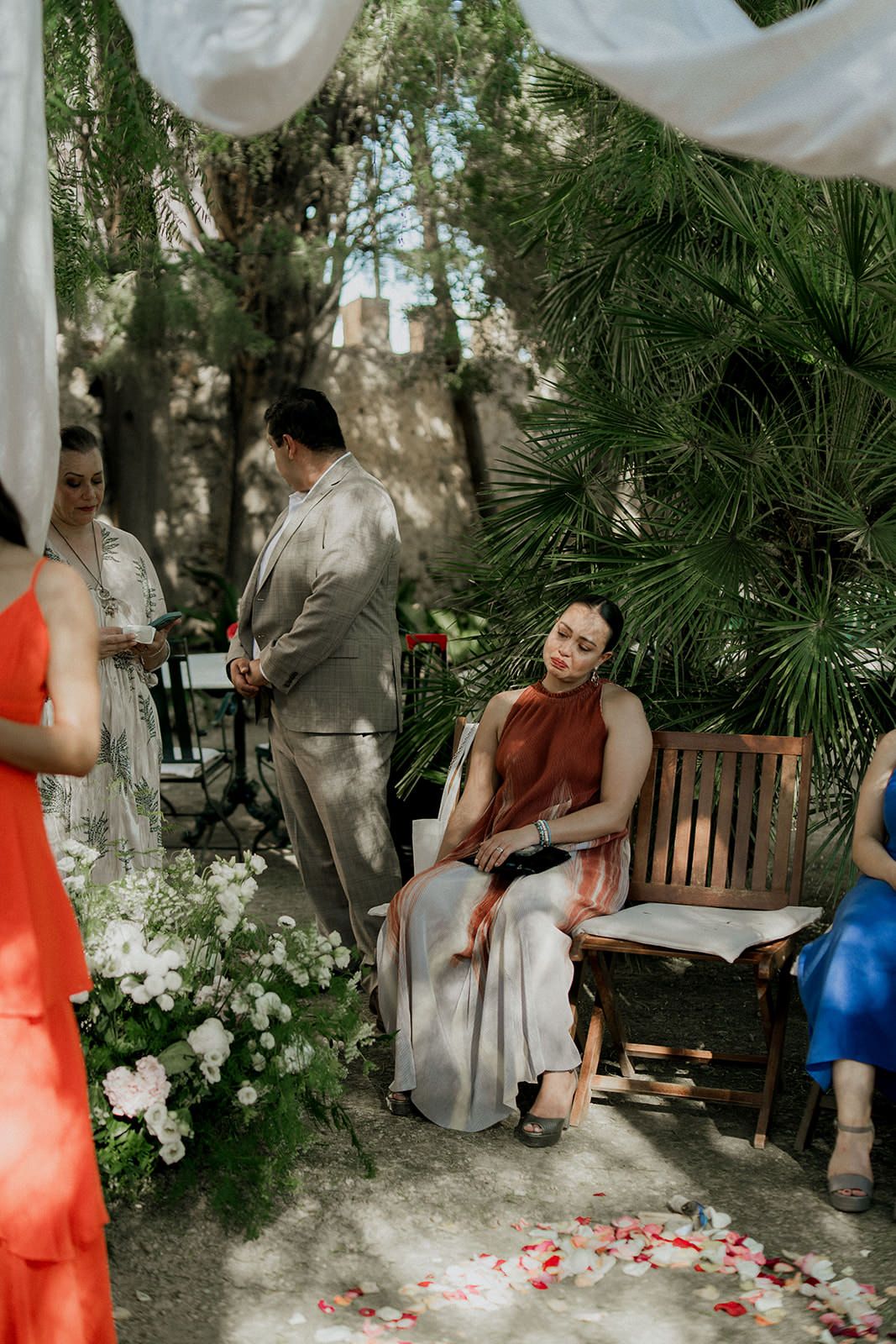 A garden wedding reception area decorated with white drapes, palm trees and scattered flower petals.