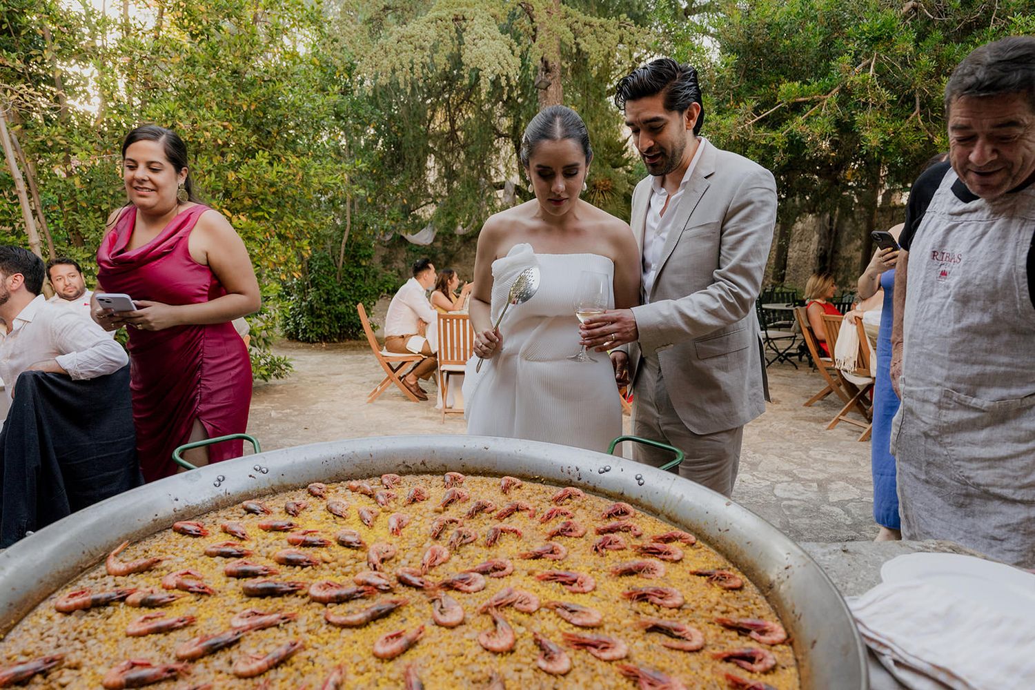 A large paella dish being prepared outdoors with people gathered around adding ingredients.