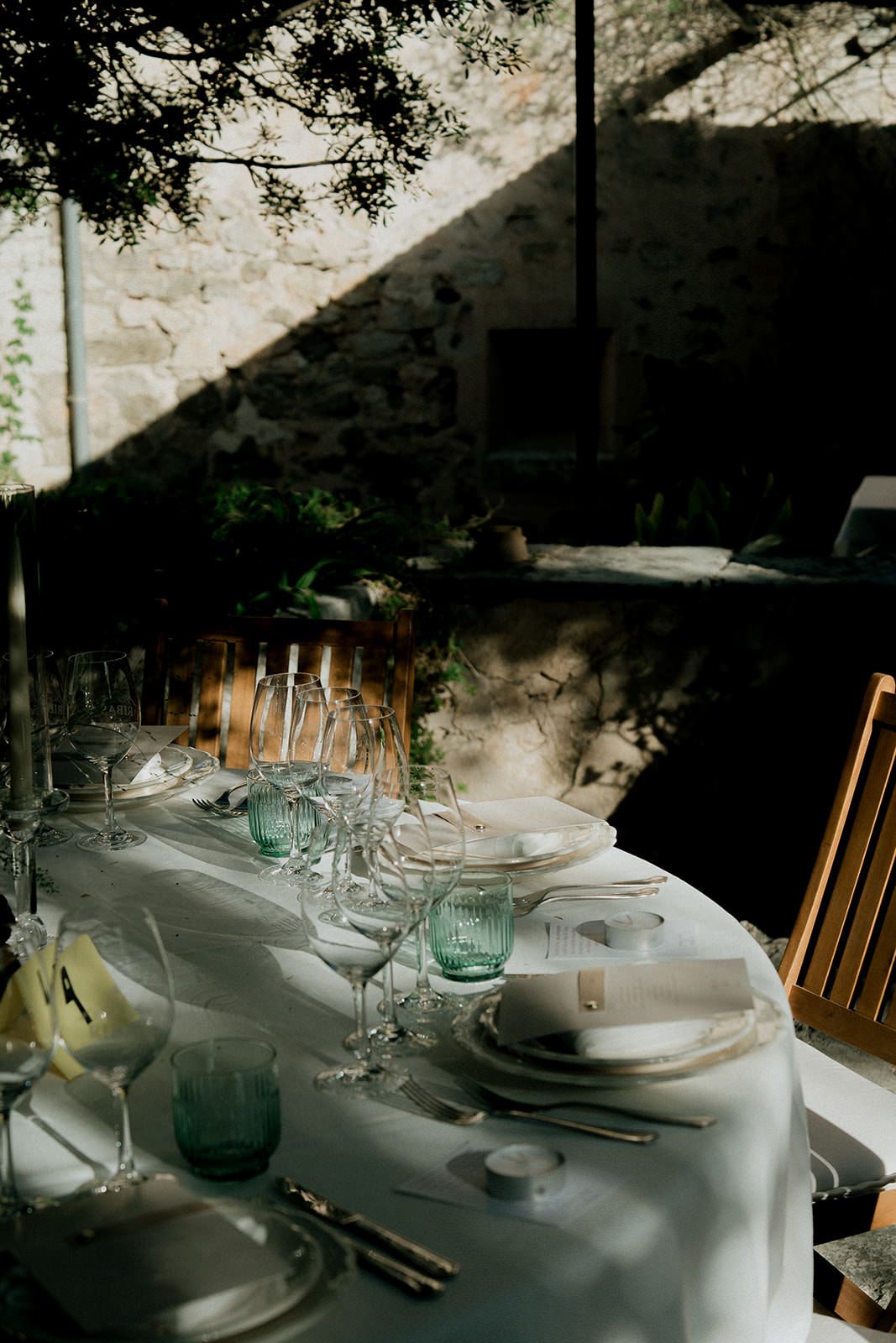 An elegant outdoor table setting with white linens and glassware under dappled sunlight and tree branches.
