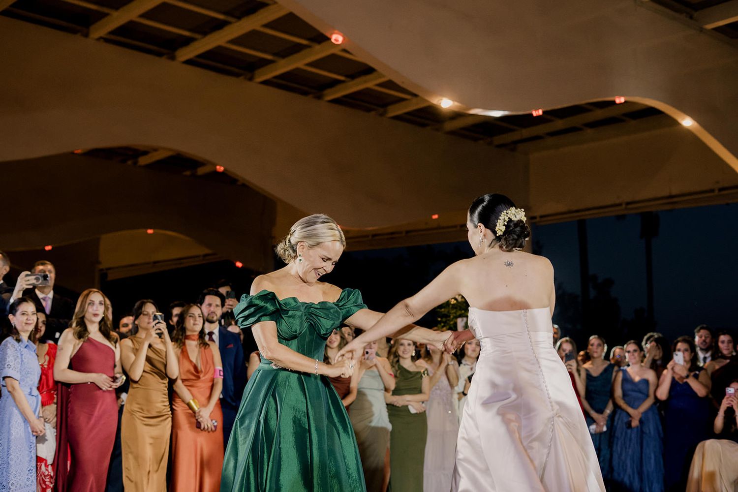 Bride in white gown dances with a woman in an emerald green off-shoulder dress at a wedding reception.