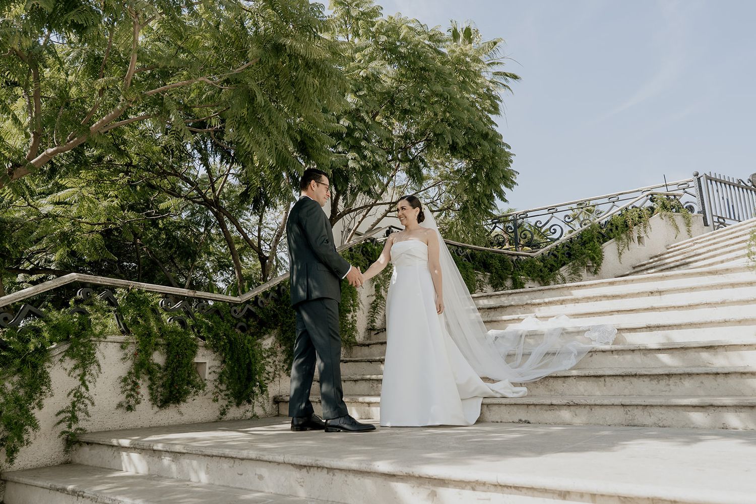 Bride and groom hold hands on elegant white stone stairs adorned with greenery on their wedding day.