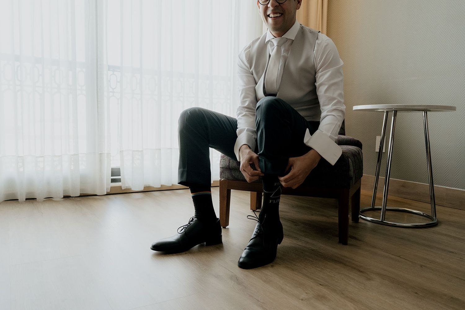 Groom sitting in a chair smiling while putting on black dress shoes before his wedding ceremony.