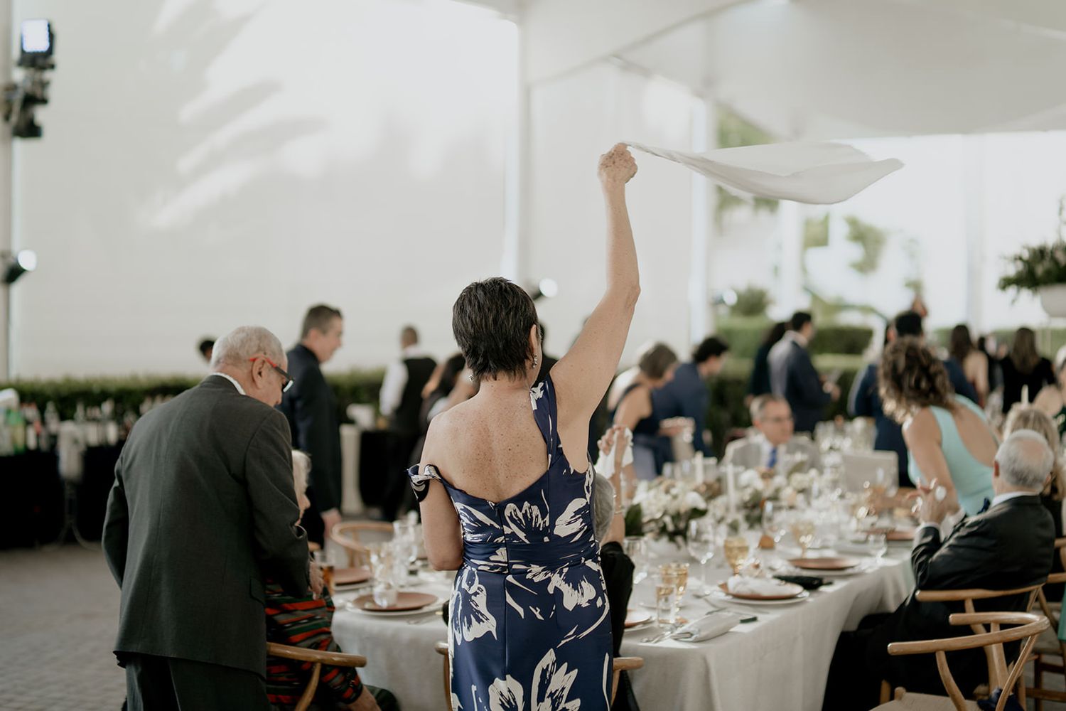 A woman in a floral dress waves a white napkin at an elegant outdoor wedding reception dinner.