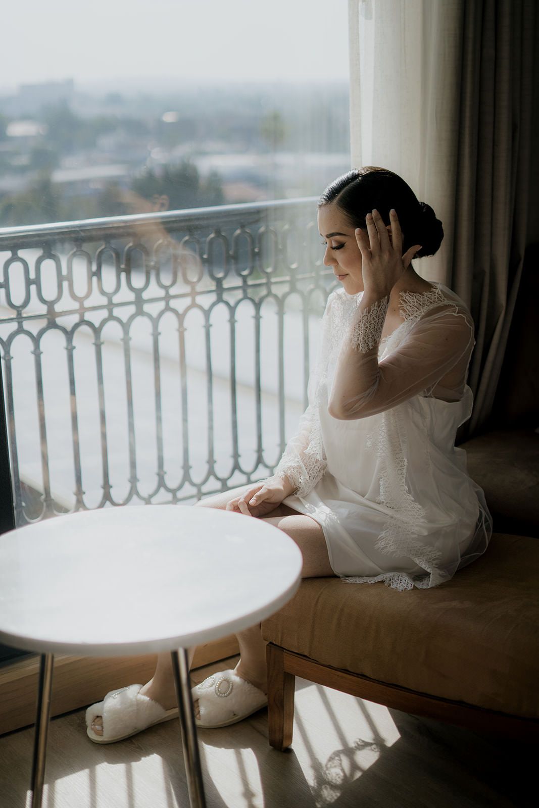 Bride in white lace robe sitting by a sunlit balcony window, adjusting her elegant updo hairstyle.