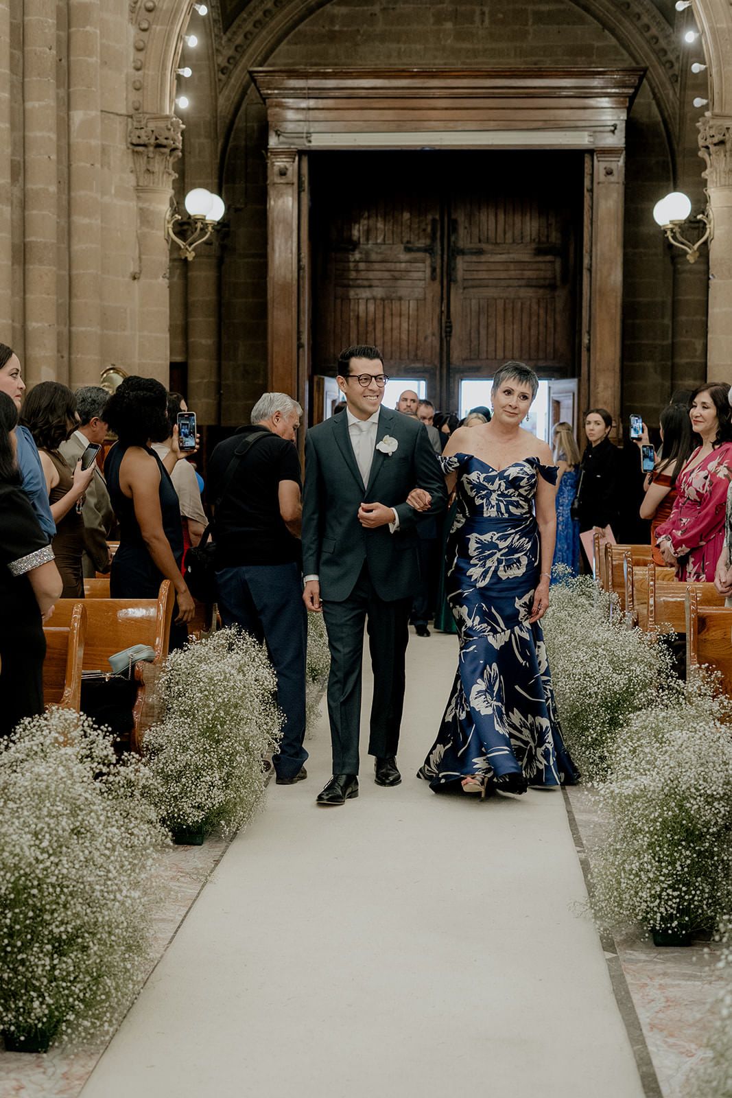 Groom in dark suit escorts woman in blue floral gown down church aisle lined with baby's breath flowers.