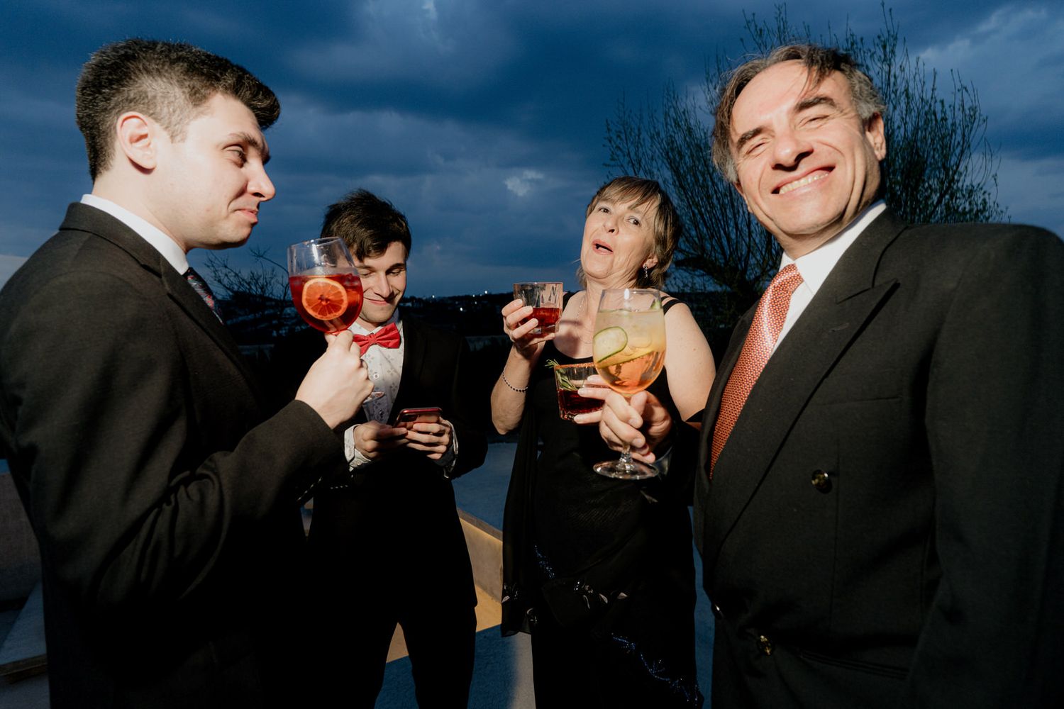 Four formally dressed people toasting with cocktails at an outdoor evening event.