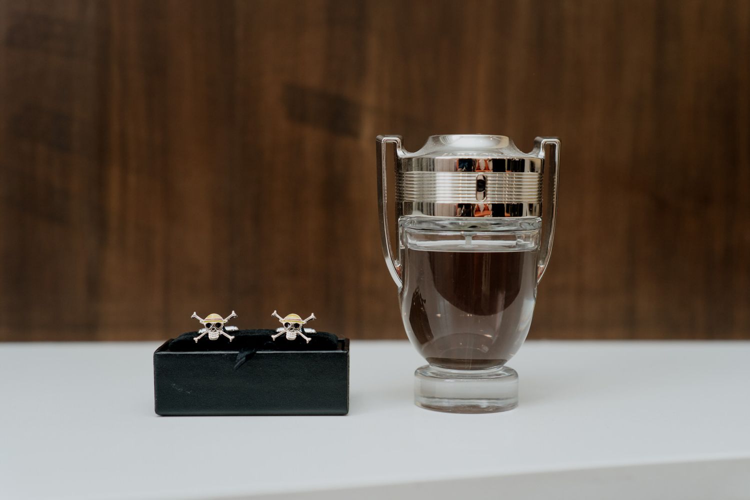 Skull cufflinks on black box beside a silver trophy-shaped cologne bottle on white surface.
