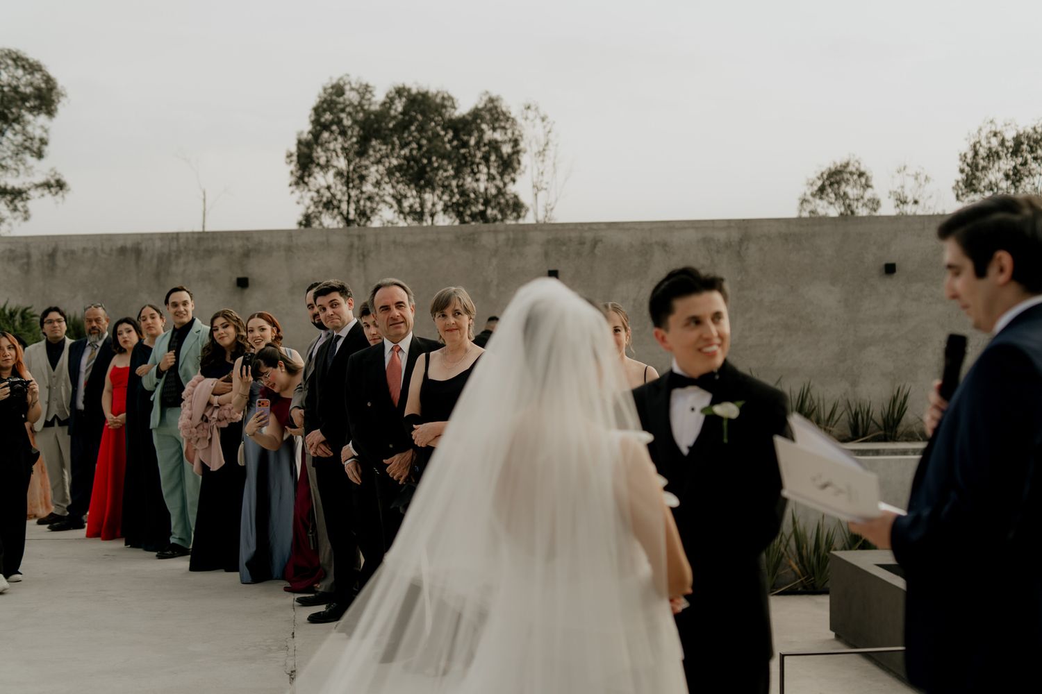 Bride in white veil walks toward groom at outdoor wedding ceremony with guests watching.