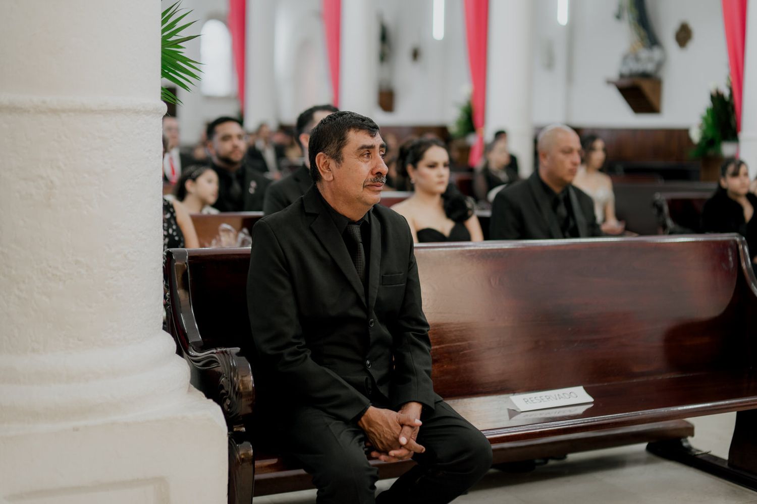 A wedding guest in formal attire sits attentively in a church pew during the ceremony.