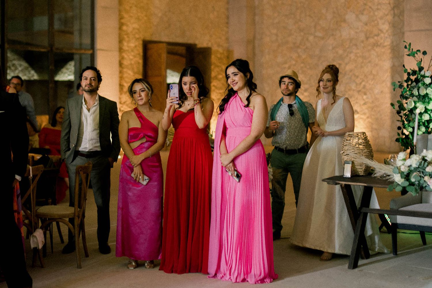 Wedding party members in pink and red dresses stand together during an indoor celebration.