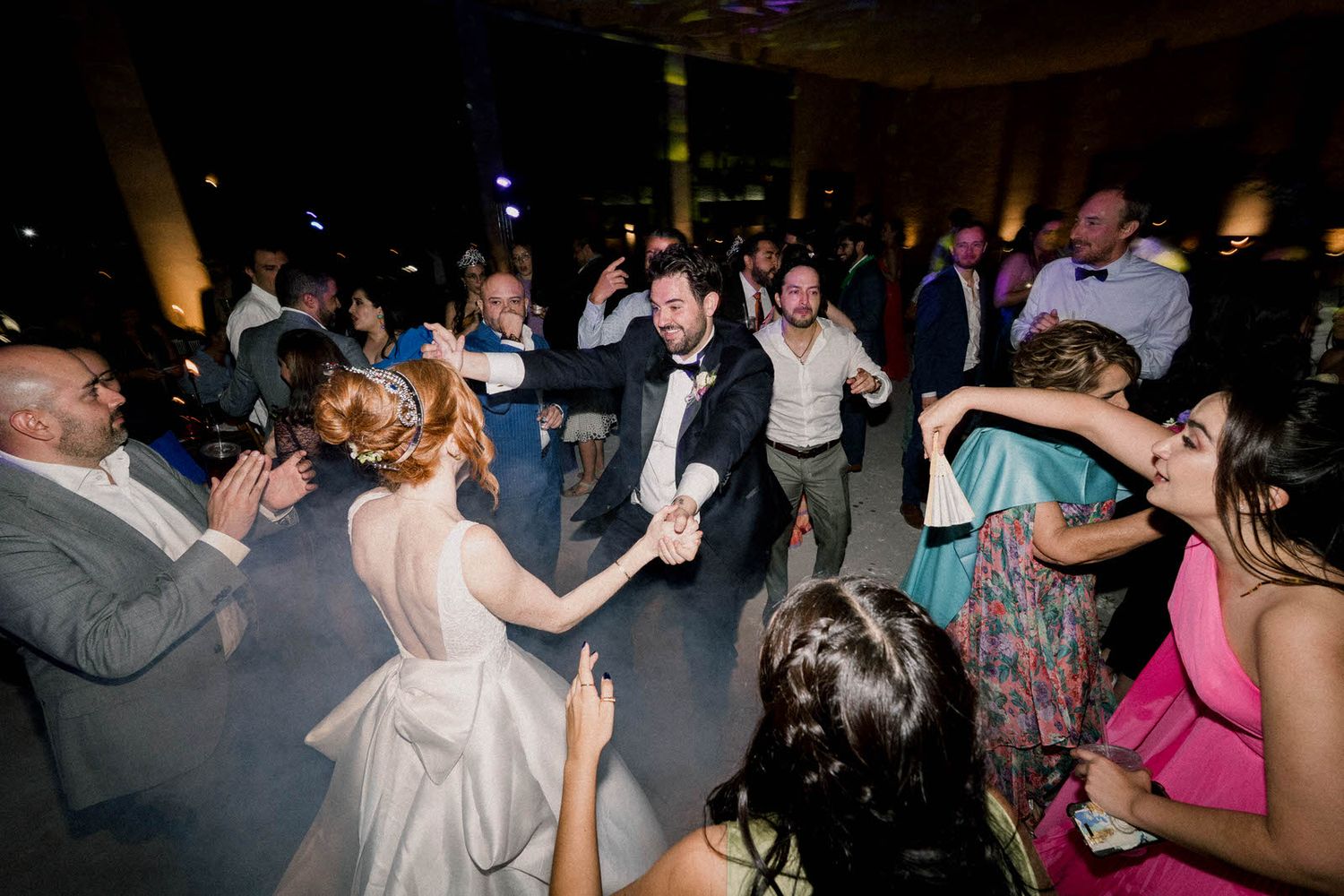 Wedding guests dance and celebrate on a crowded reception dance floor under ambient lighting.