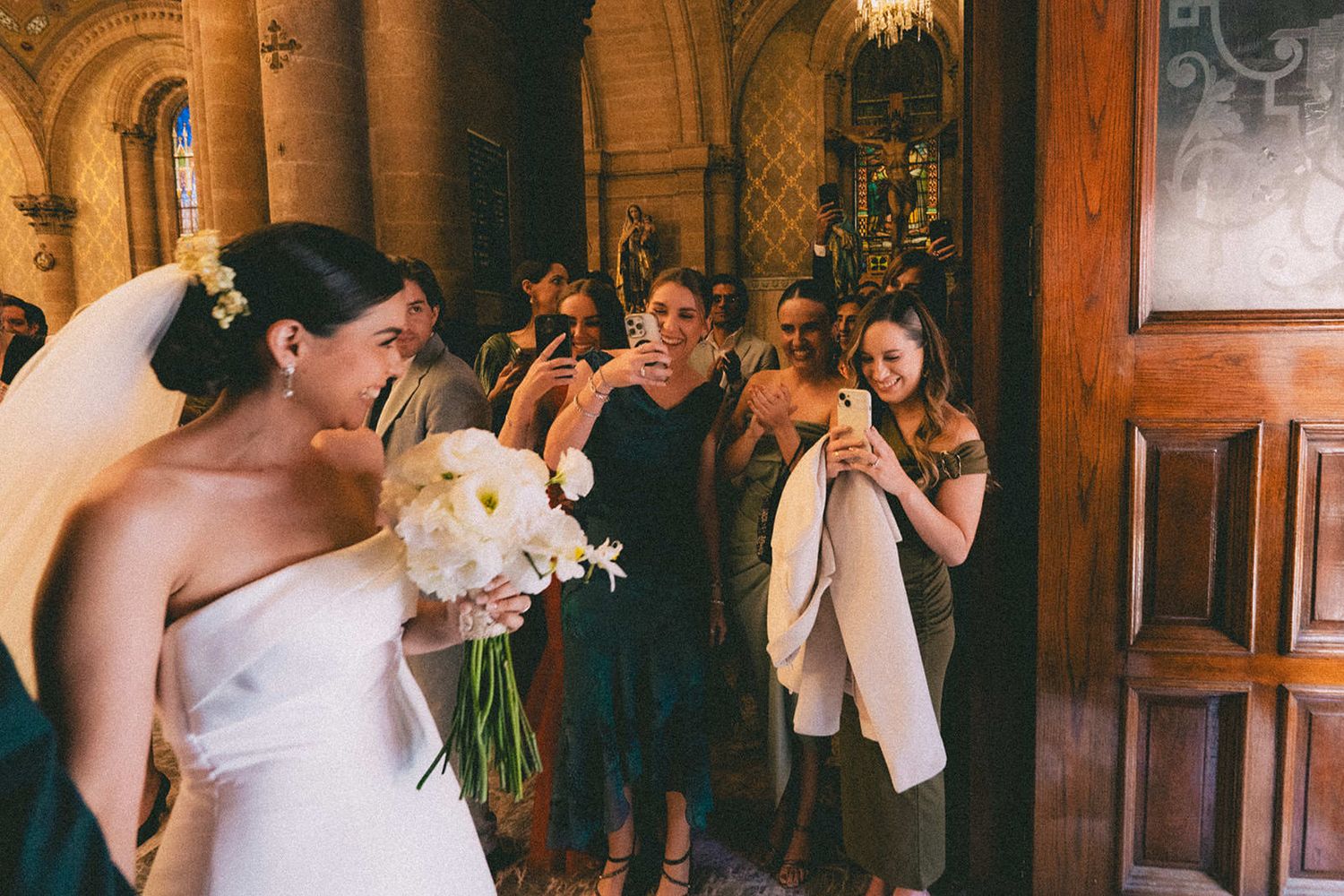 A bride in a strapless white gown holds a white bouquet while guests photograph her at a church wedding.