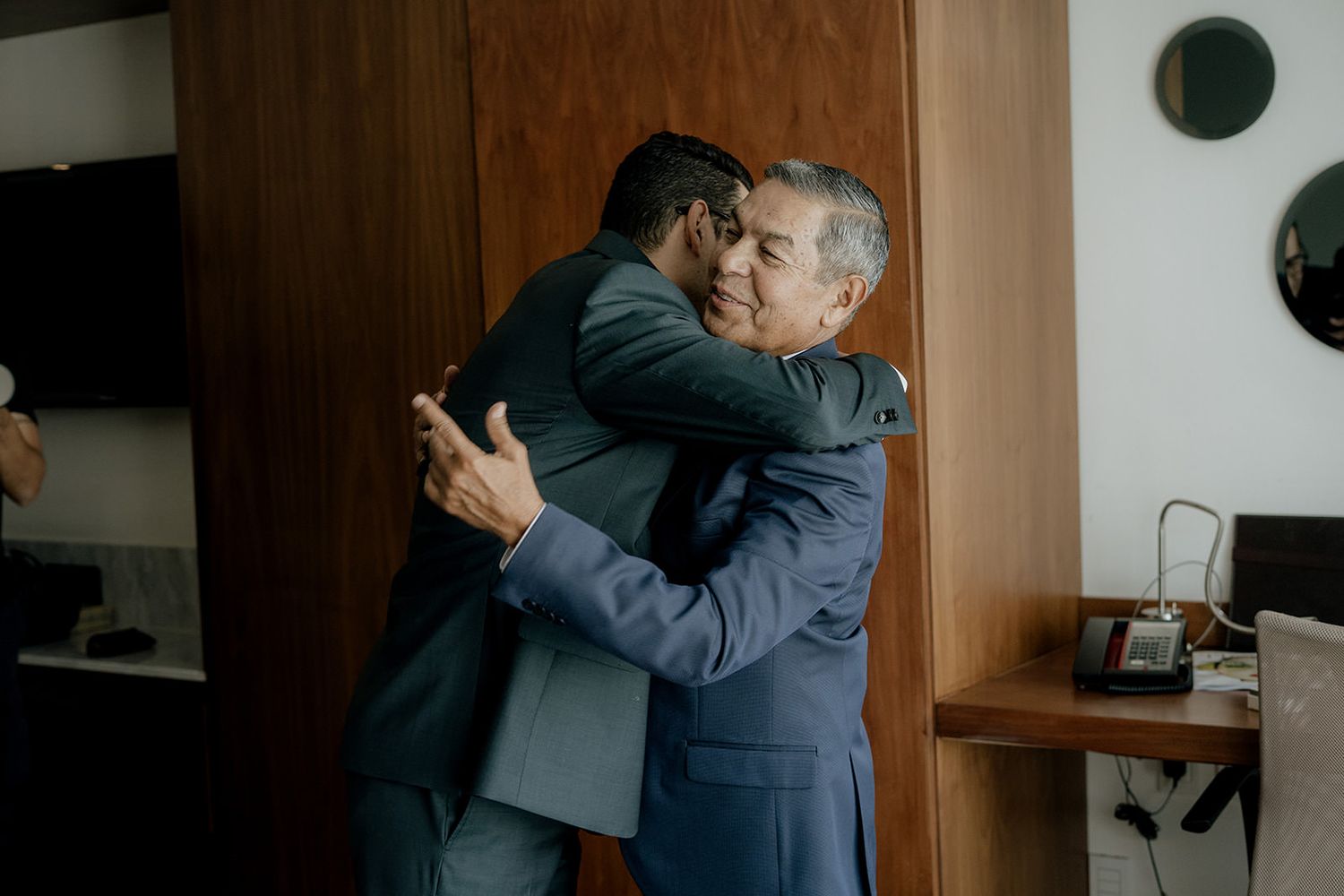 Two men in suits share a warm embrace in a hotel room during a wedding day celebration.