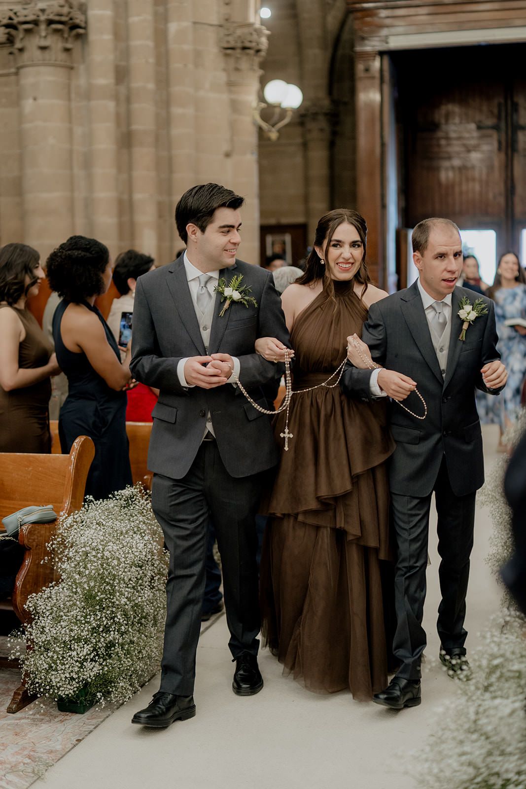 Bride in brown halter gown walks down church aisle escorted by two groomsmen holding rosary at wedding.