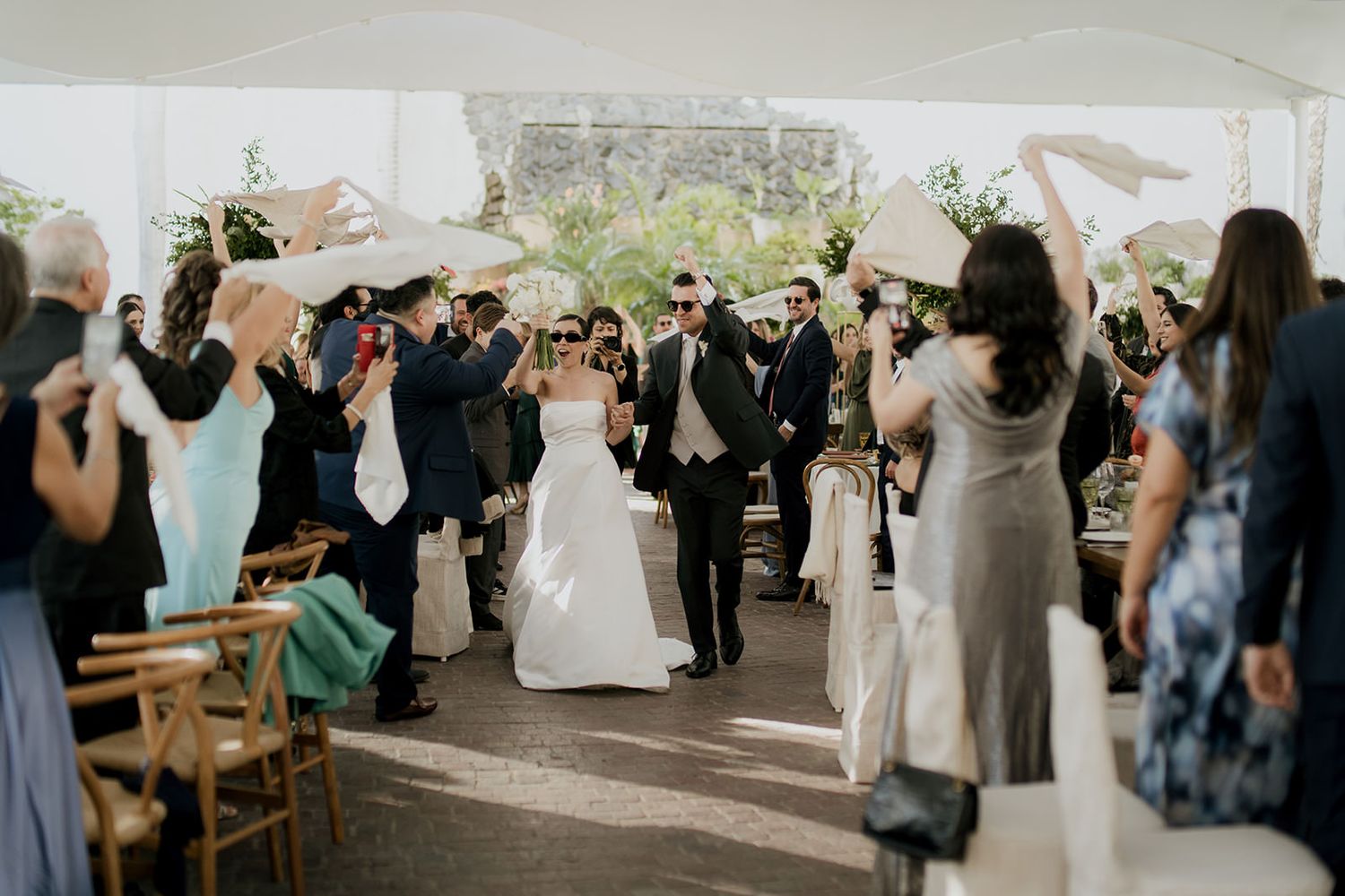 Joyful newlyweds walk down aisle as guests wave fans during outdoor wedding ceremony under white tent.