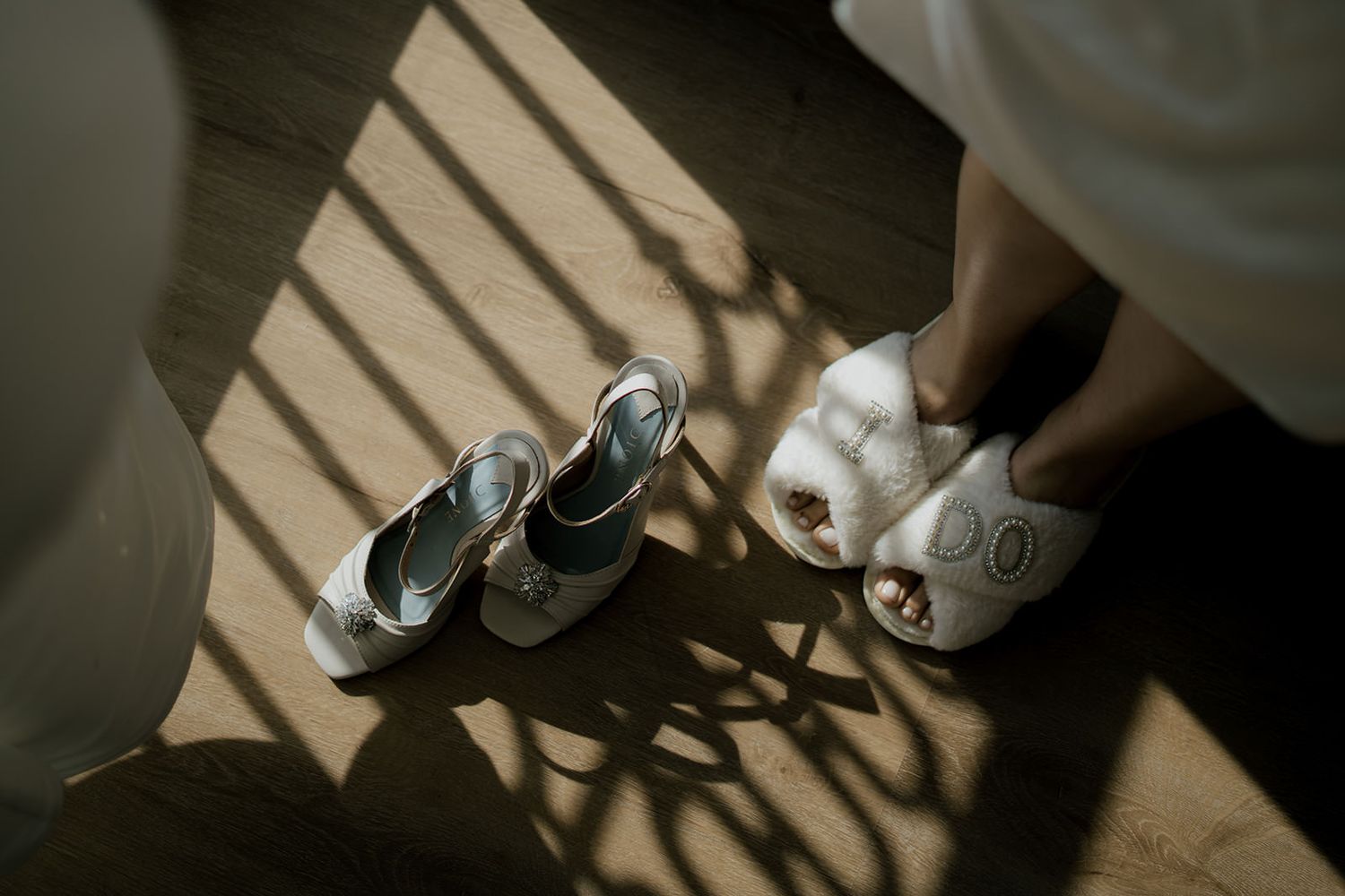 Two pairs of white bridal heels on a wooden floor with dramatic sunlight shadows, one pair being worn by a bride.