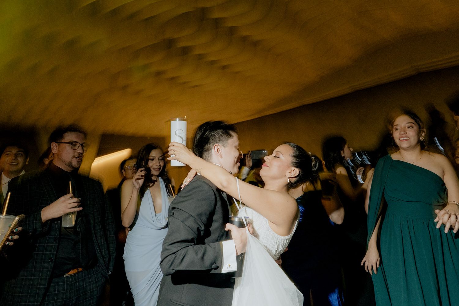 Bride and groom share a joyful kiss on the dance floor surrounded by celebrating wedding guests.