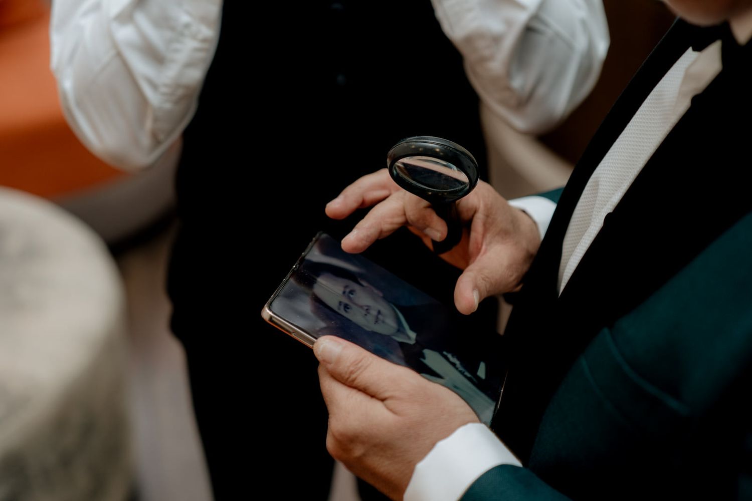 Groom in tuxedo examines vintage photo with magnifying glass at wedding reception.