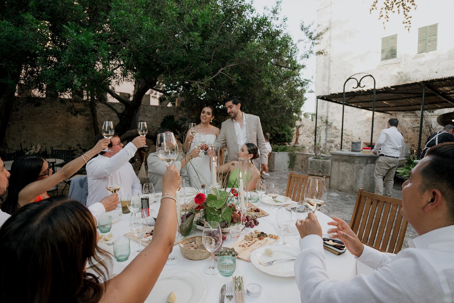 Guests seated at an elegant outdoor wedding reception with white table settings and natural greenery overhead.