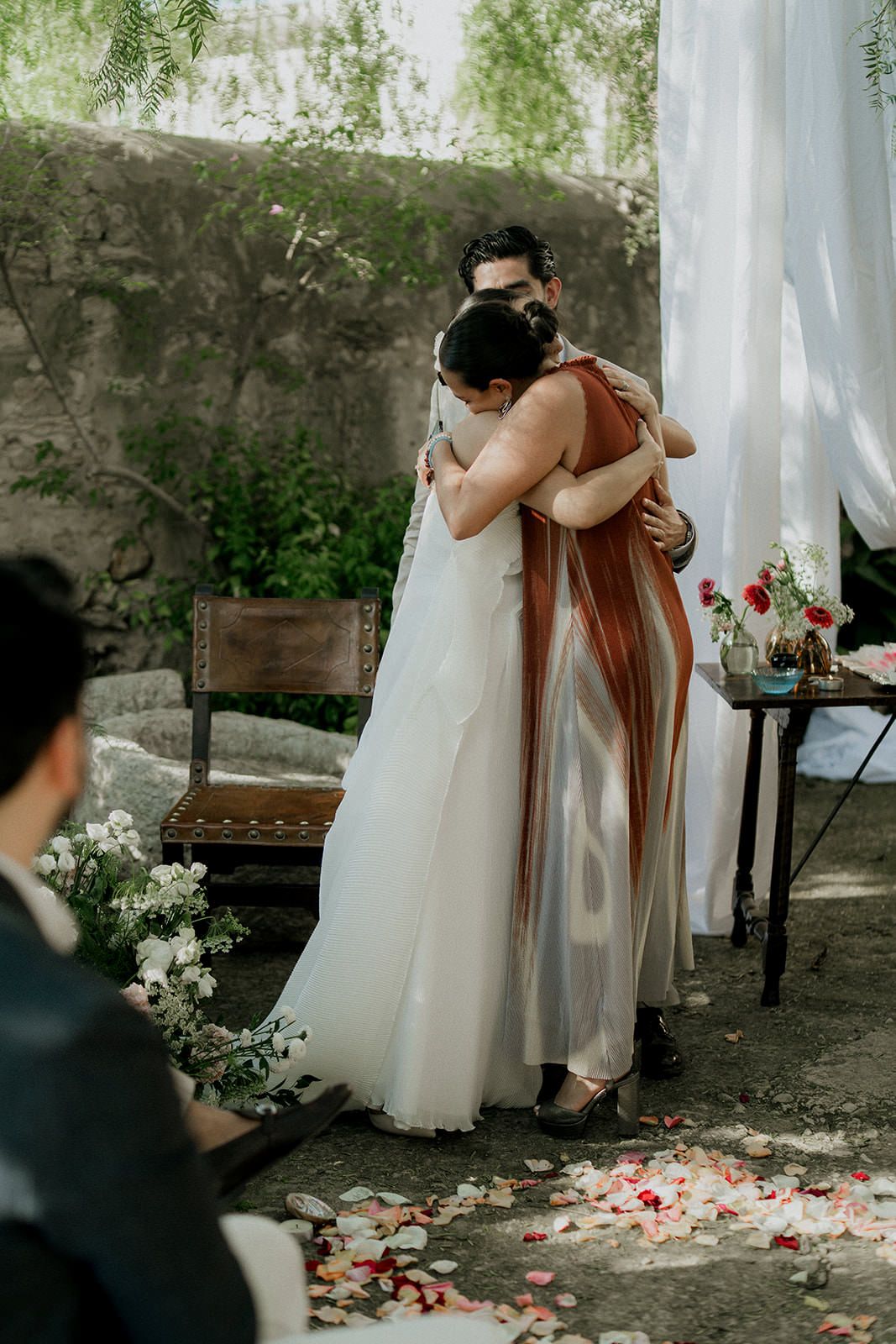 Two people share an emotional embrace at an outdoor wedding ceremony with scattered rose petals on the ground.