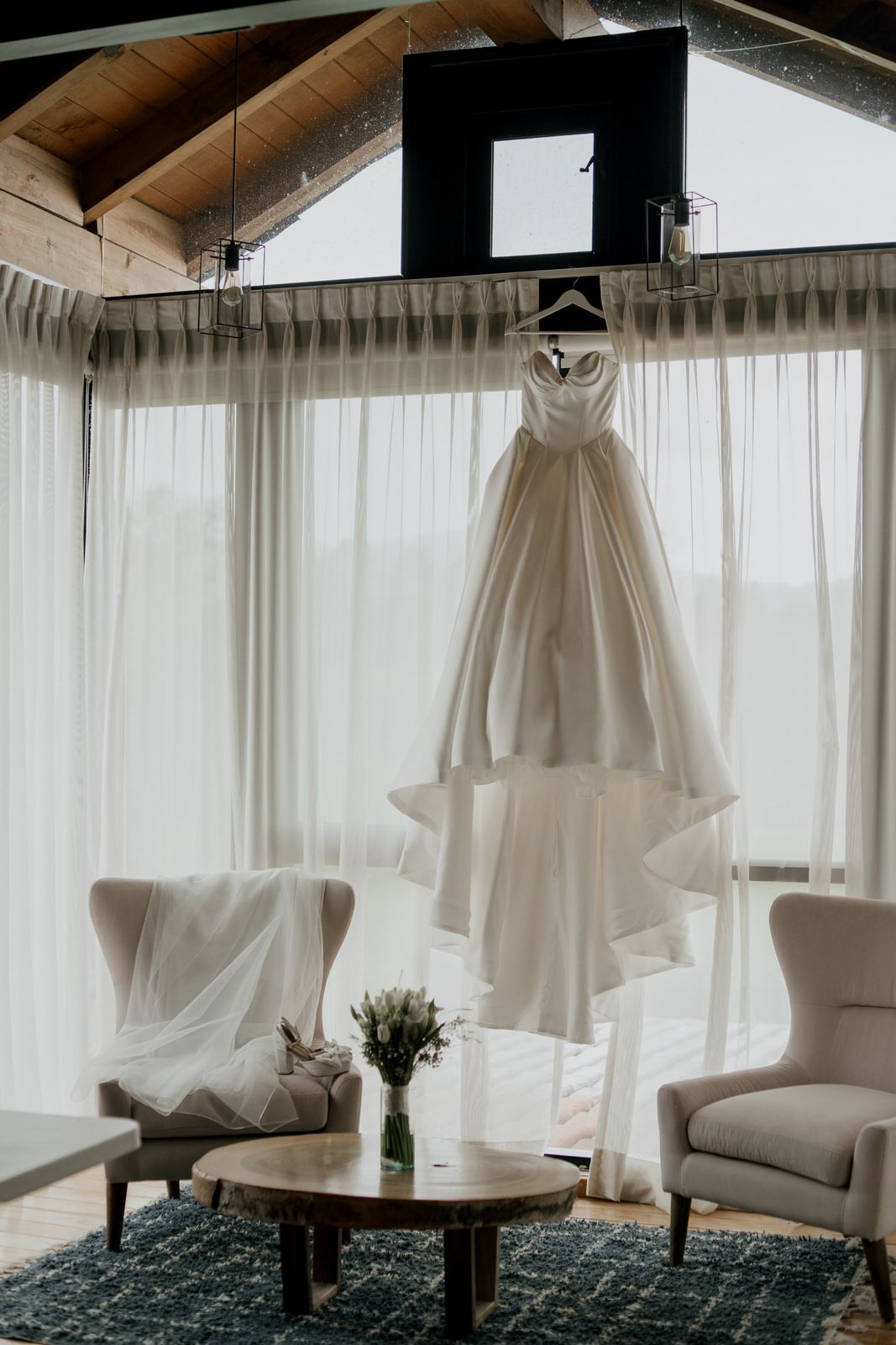 White wedding dress hanging by window in bright modern room with blue rug and armchairs.