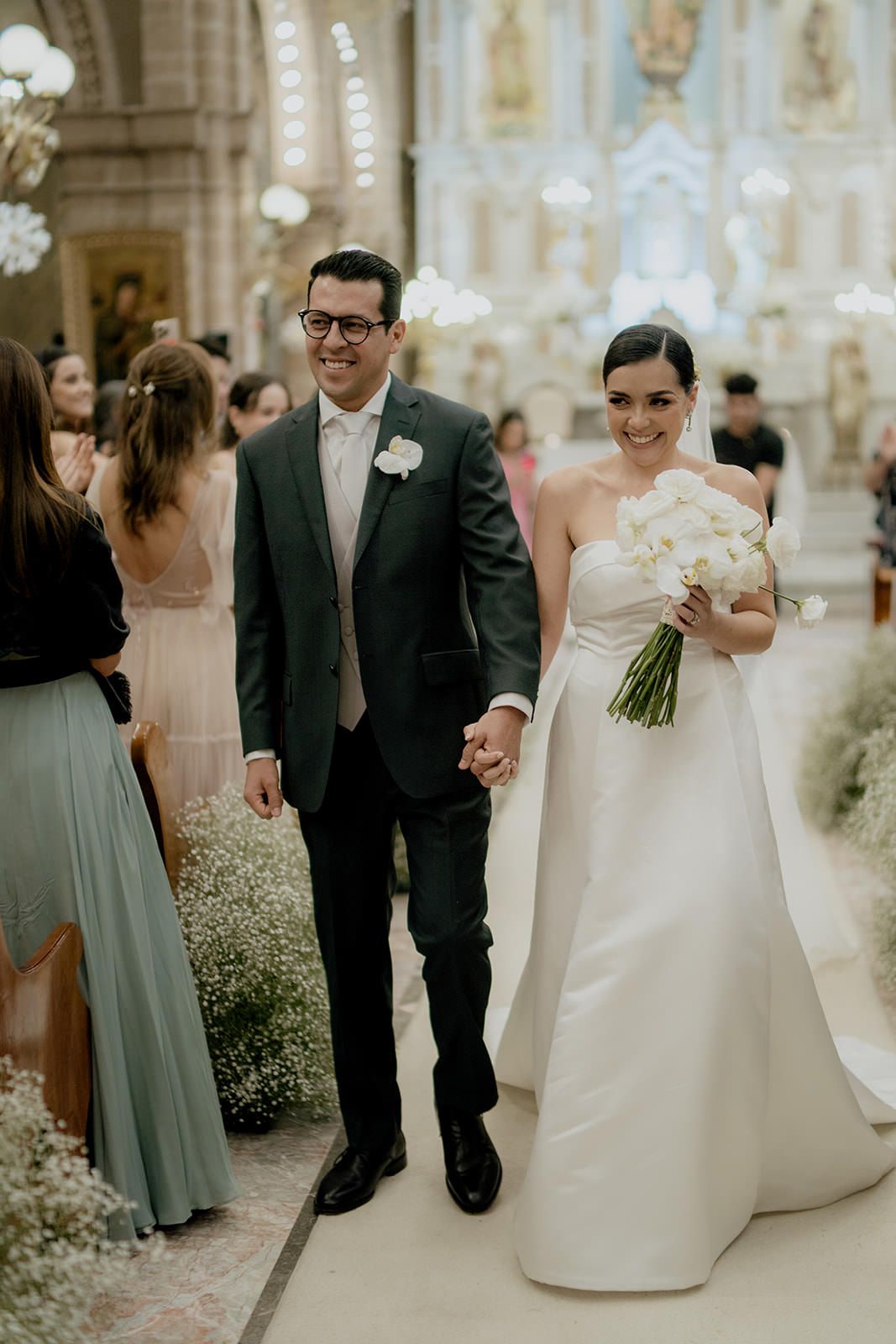 Smiling newlyweds walk down church aisle, bride in strapless gown holding white bouquet, groom in dark suit.