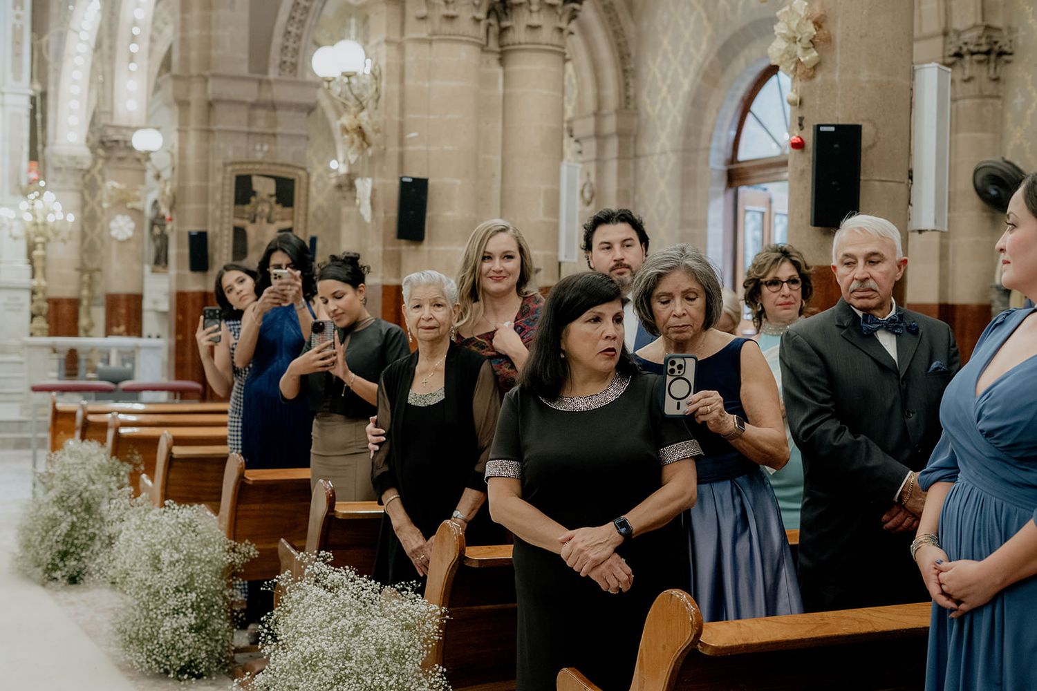 Wedding guests standing in church pews, watching ceremony aisle decorated with baby's breath floral arrangements.