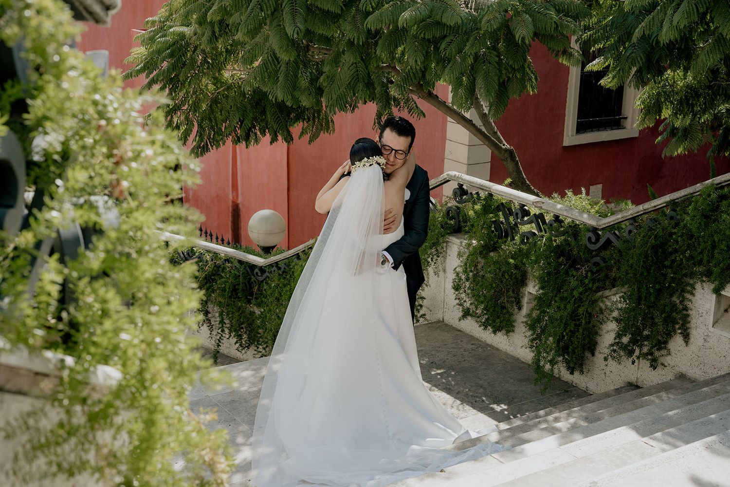 Bride and groom share a romantic embrace on outdoor stairs surrounded by lush greenery and a red building.