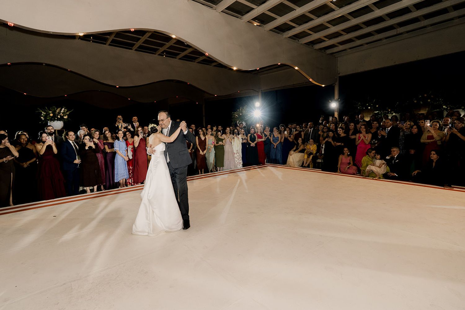 Bride and groom share first dance on white dance floor surrounded by wedding guests at night.